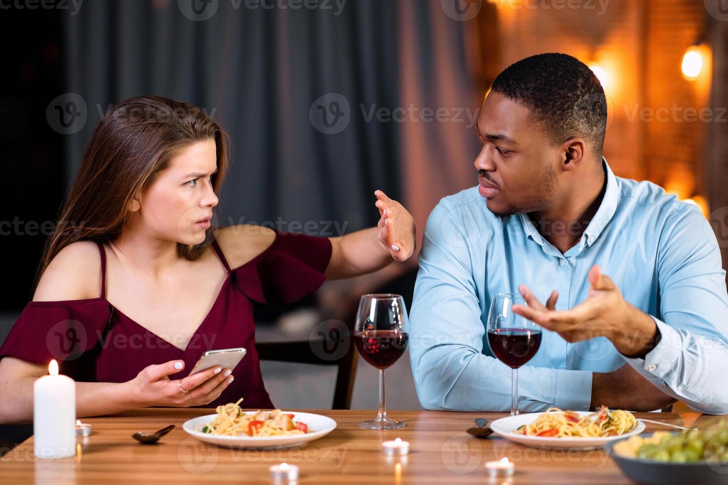 Annoyed Black Guy Blaming His Girlfriend For Using Smartphone During Romantic Dinner In Restaurant, Young Woman Showing Stop Gesture With Hand, Multicultural Couple Having Relationship Problems photo