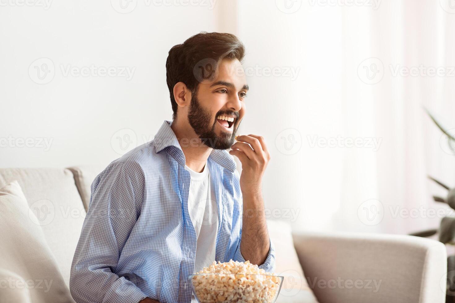 Cheerful eastern man spending free time at home, watching TV and eating popcorn. Happy arab guy enjoying comedy movie while relaxing on couch in living room, side view with free space photo