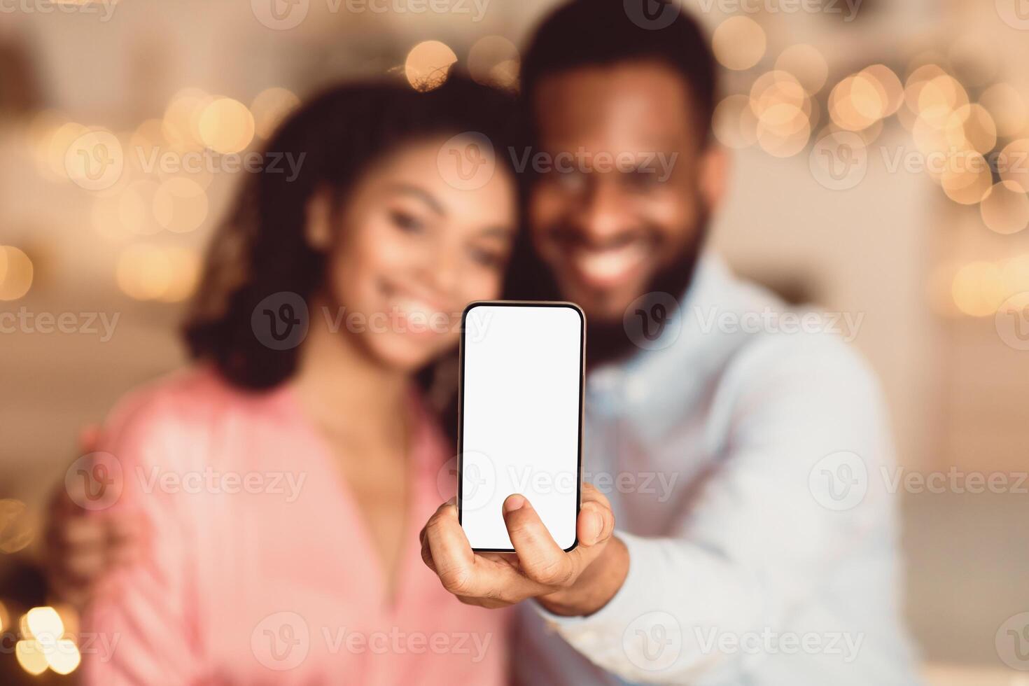 People And Technology Concept. Smiling African American Man Holding And Showing Smartphone With White Blank Empty Screen For Mockup, Hugging Woman In Blurred Background, Selective Focus photo