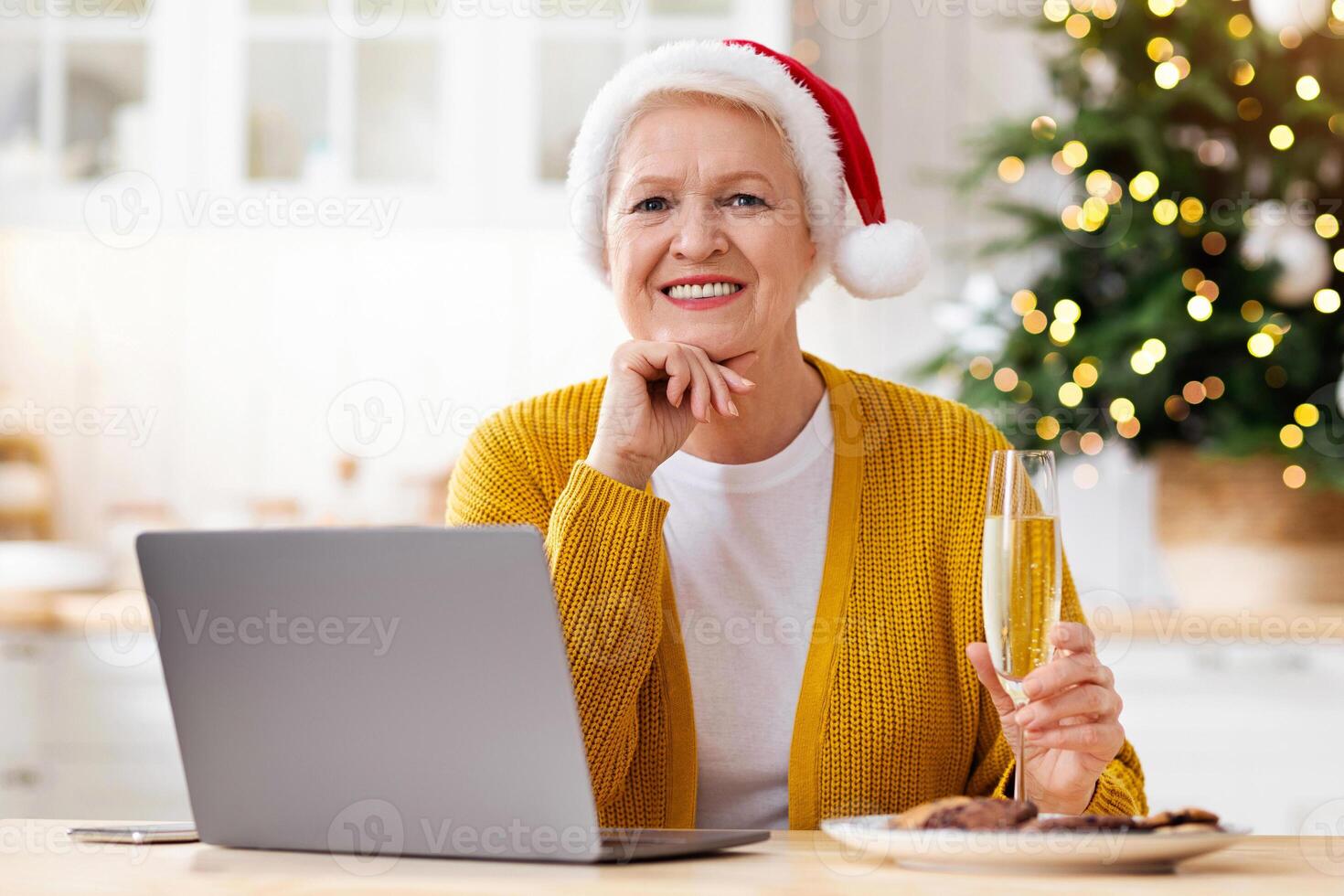 Beautiful granny in Santa hat sitting in front of laptop at kitchen, having Chritsmas tree behind, holding glass of champagne and smiling at camera, empty space. Old woman celebrating New Year alone photo