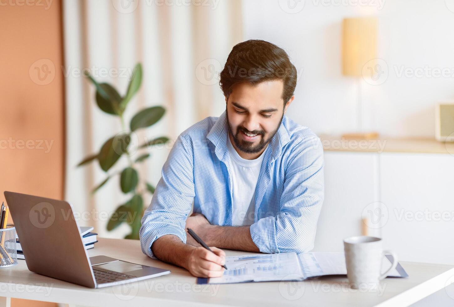 Smiling Arab Man Working With Documents And Laptop At Home Office, Handsome Eastern Male Entrepreneur Sitting At Desk With Computer, Checking Financial Reports And Taking Notes, Free Space photo
