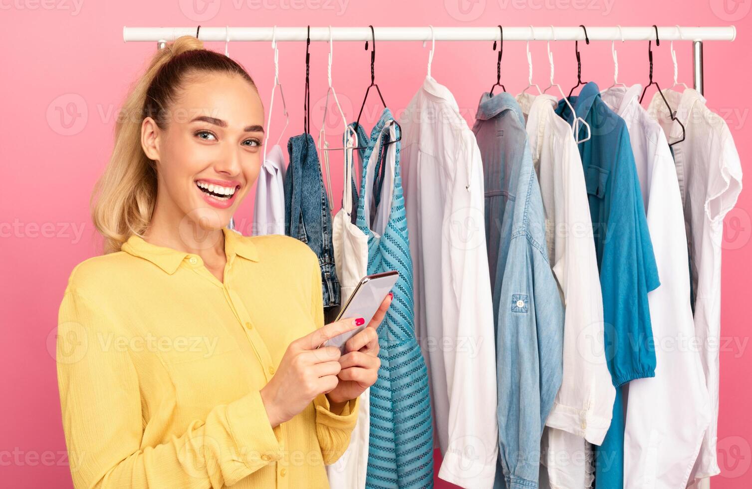 Season Of Sales And Discounts. Portrait of smiling young woman holding and using her mobile phone, chatting online or making purchases via internet standing near clothing rail at the wardrobe photo