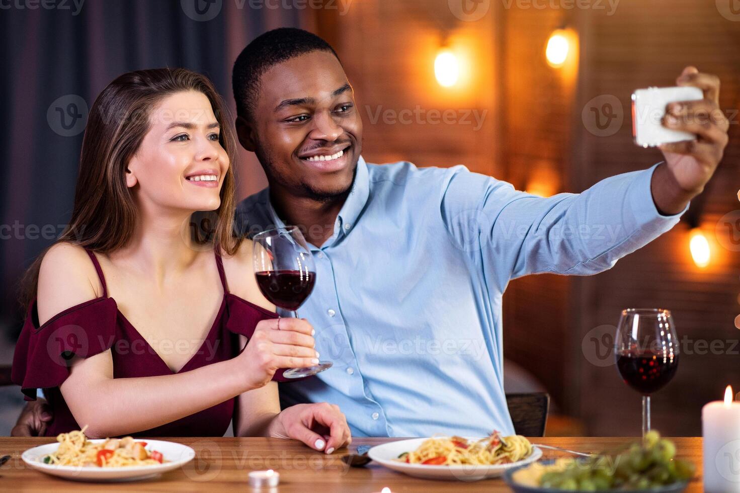 Happy Interracial Couple Taking Selfie On Smartphone During Romantic Date In Restaurant. Cheerful Black Man And White Woman In Love Smiling At Phone Camera, Celebrating Valentine's Day Together photo