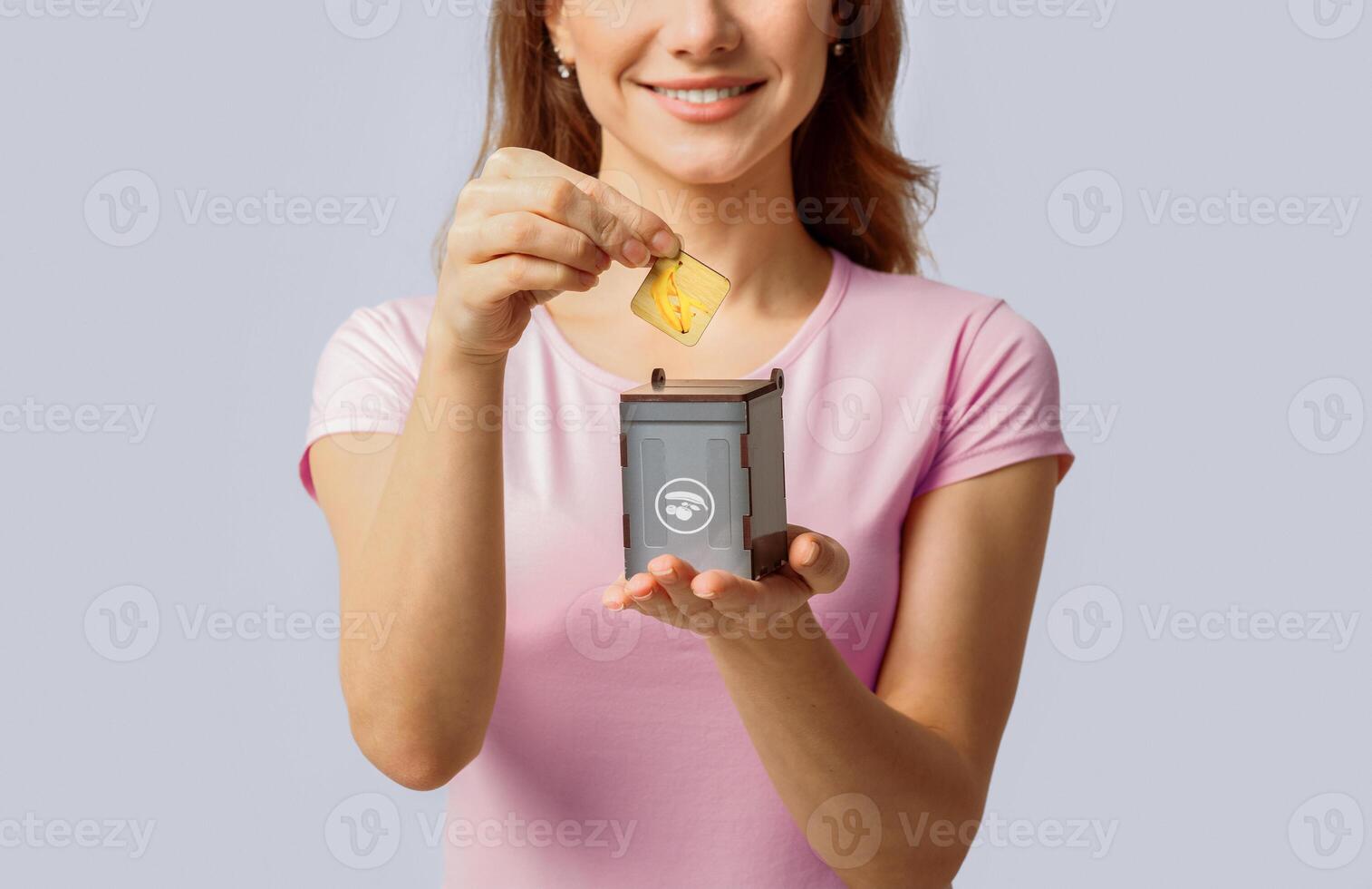 A millennial woman happily participates in recycling by placing a yellow item into a small recycling bin. She showcases her commitment to eco-friendly practices in a minimalistic background. photo