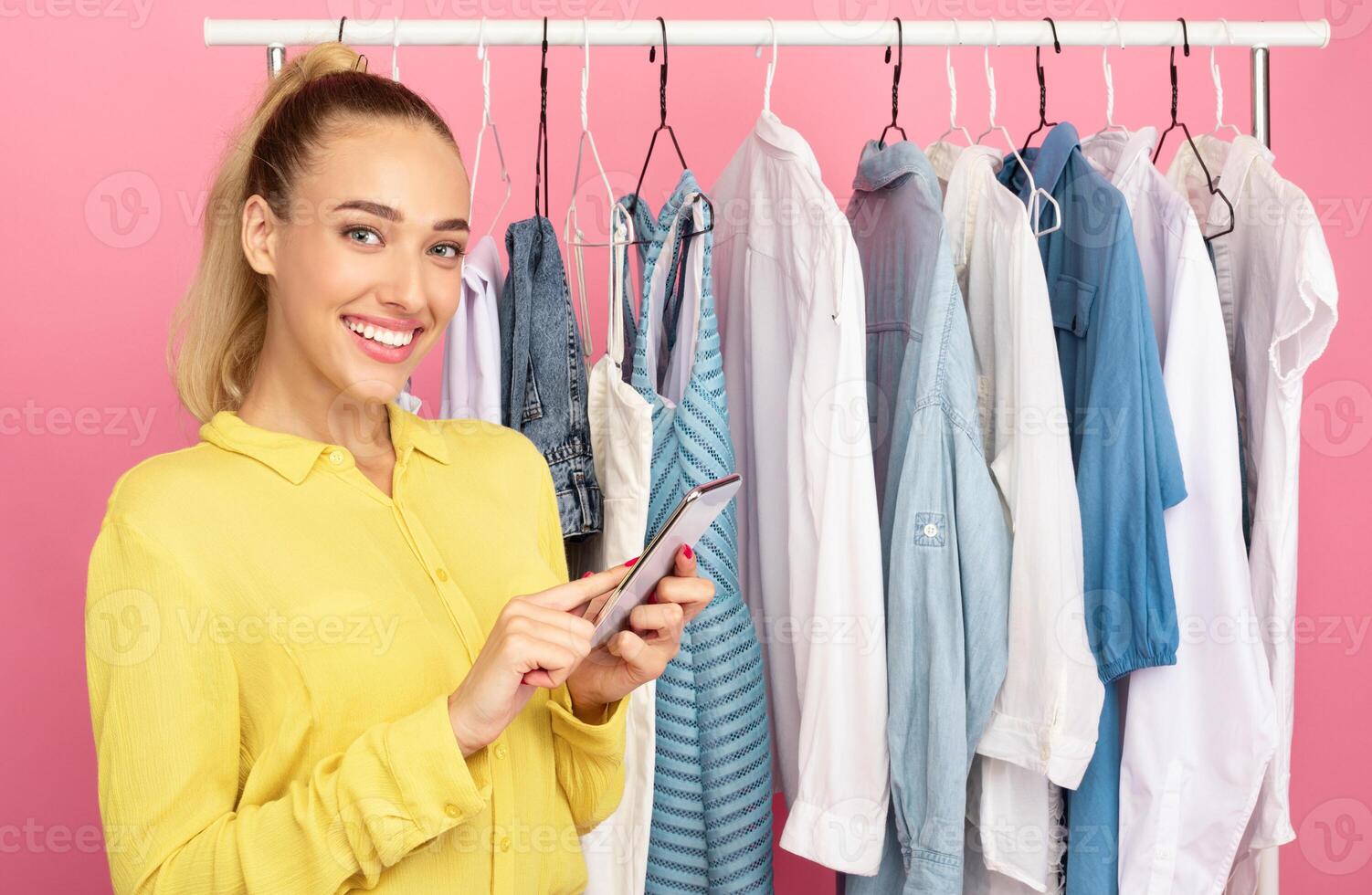 Fashion And Style. Portrait of smiling woman stylist holding and using smartphone, texting sms or making purchases in online store standing near rack with clothes over pastel pink studio background photo