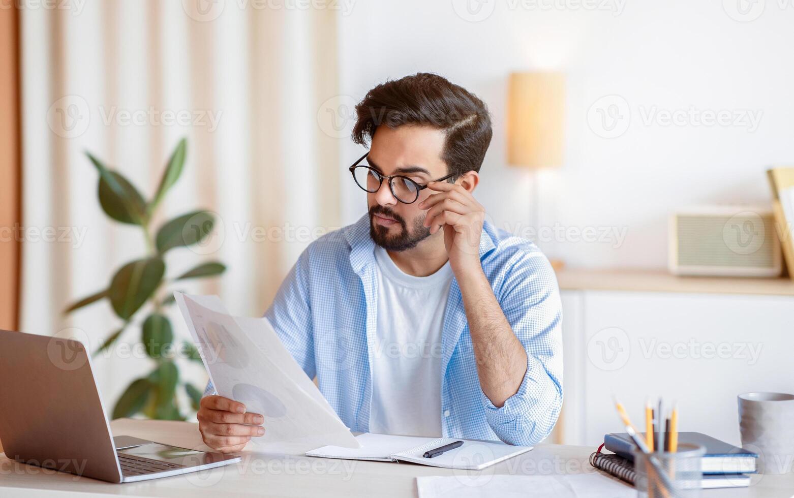 Young Eastern Businessman In Eyeglasses Working With Papers And Laptop In Office, Millennial Arab Entrepreneur Reading Reading Financial Documents While Sitting At Worplace, Free Space photo