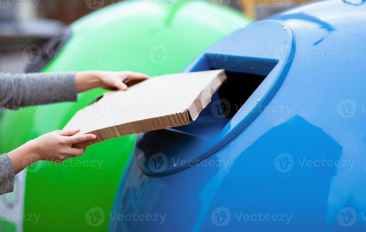 Waste Sorting Concept. Hand throwing cardboard box into blue recycle bin container outdoors, unrecognizable woman enjoying garbage recycling system, cropped image with selective focus photo