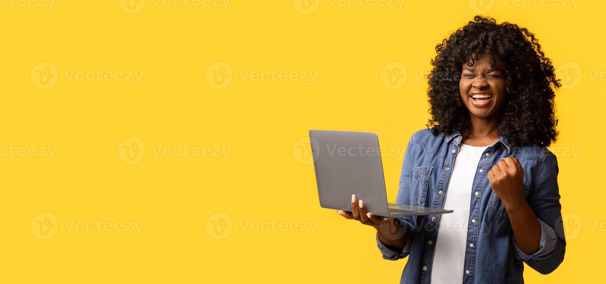 Emotional african american young woman with laptop celebrating success, gambling online, raising fist up and screaming over yellow studio background, panorama with empty space photo