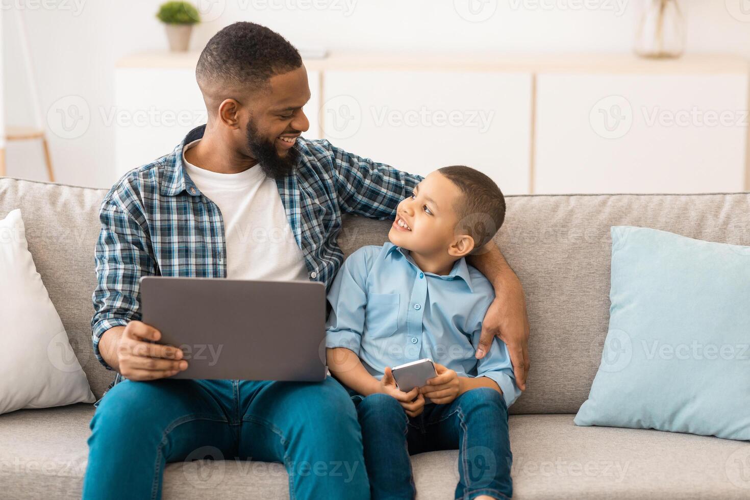 African Daddy And Little Son Using Digital Tablet Computer Browsing Internet Together At Home, Sitting On Sofa Indoors. Gadgets And Family Weekend. Father And Kid Watching Movie Online photo