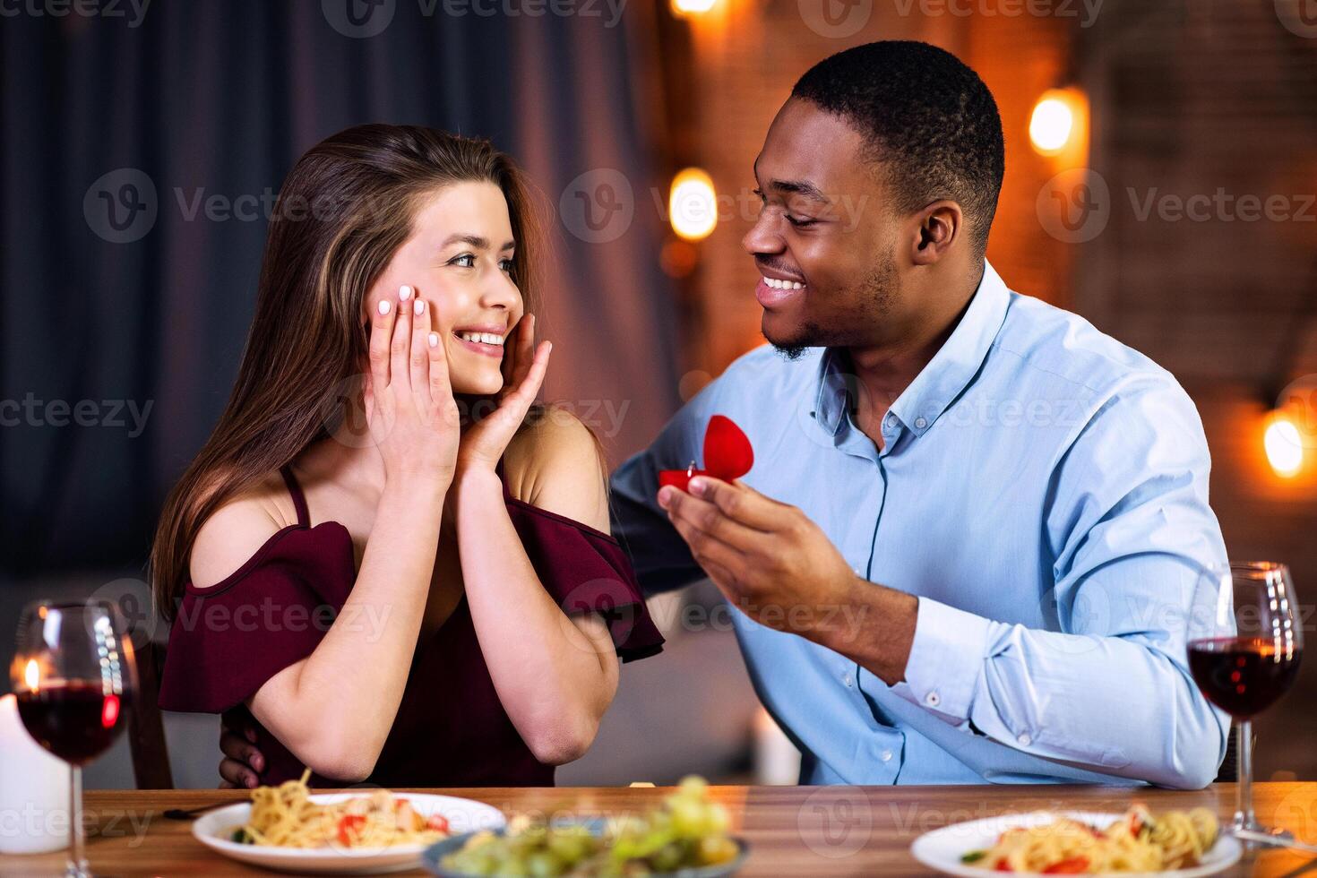 Happy African American Man Making Proposal To His Surprised White Girlfriend, Asking To Marry Him During Romantic Date Dinner In Restaurant, Giving Box With Diamond Ring, Free Space photo