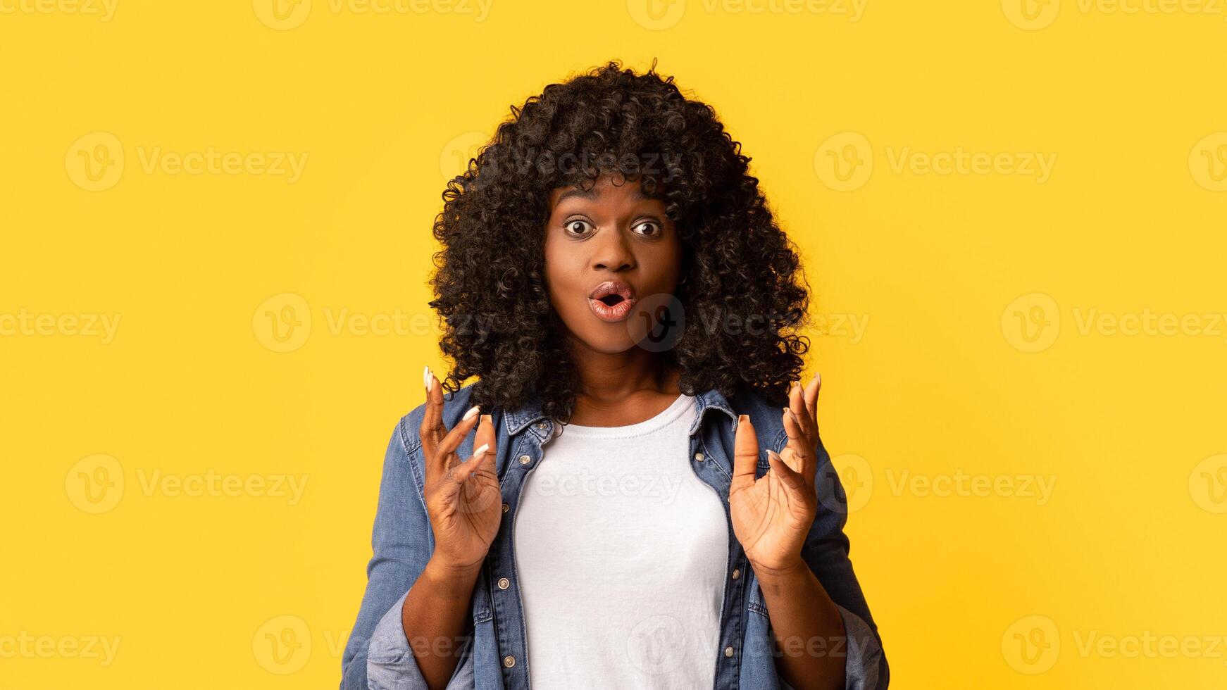 Amazed african american curly young woman with eyes and mouth wide open raising hands up over yellow studio background, seeing something shocking, copy space. Emotions, face expression concept photo
