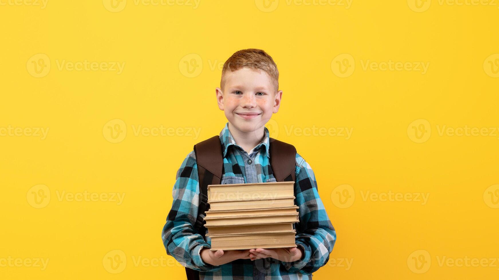 Smiling schoolboy with backpack holding heap of books, yellow studio background, copy space. Happy boy going to school, holding books, enjoying his school days. Kids education concept photo