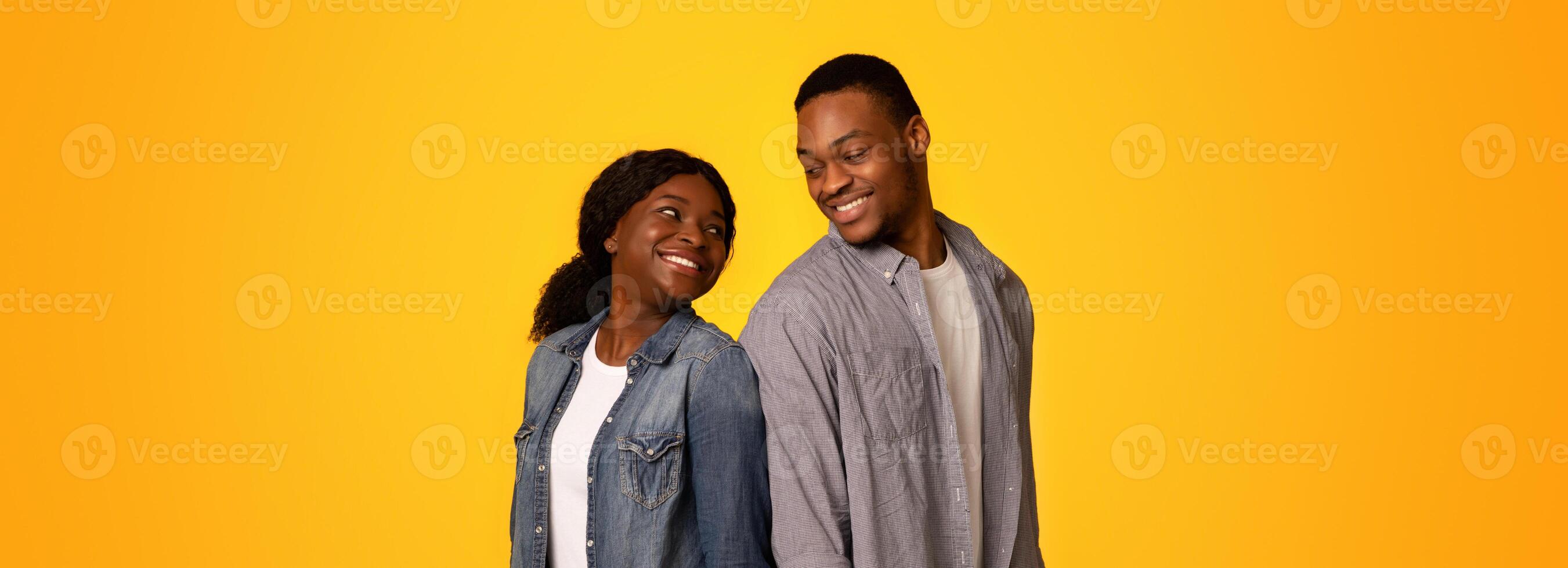Cheerful black man and woman standing back to back and turning heads to each other while standing isolated over yellow background, young african american lovers smiling and having fun, copy space photo