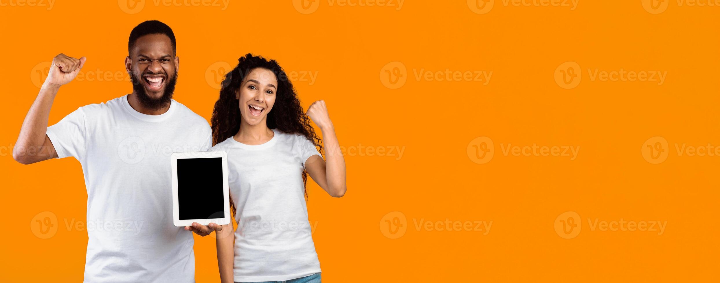 Diverse Couple Showing Tablet Empty Screen Shaking Fists Celebrating Good News Standing Over Yellow Studio Background, Smiling To Camera. Great Mobile Application. Mockup, Panorama With Blank Space photo