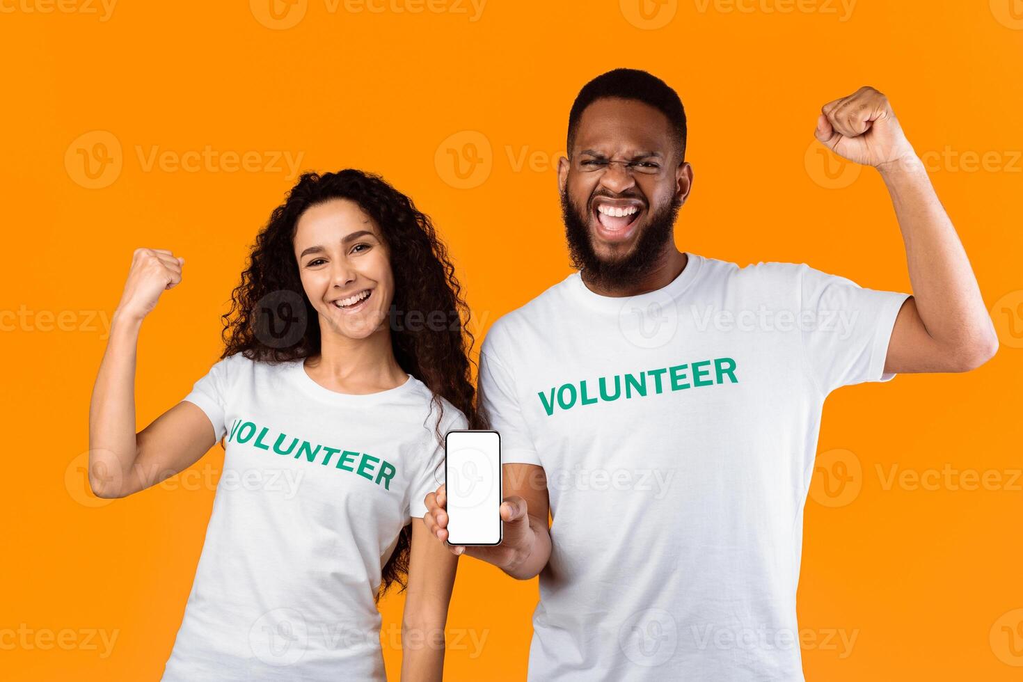 Volunteering. Two Diverse Volunteers Showing Smartphone With Blank Screen Recommending Volunteering App Standing On Yellow Studio Background. Power Of Kindness Concept. Mockup photo