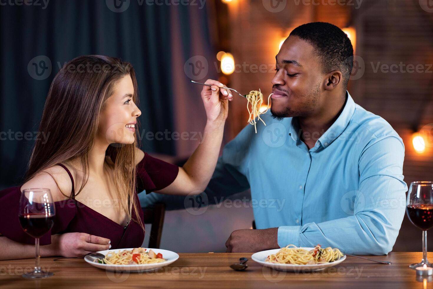 Cheerful Interracial Couple Having Fun During Romantic Dinner At Home. Young Multiracial Lovers Eating Spaghetti And Drinking Wine, Caring Woman Feeding Her Boyfriend And Smiling, Free Space photo