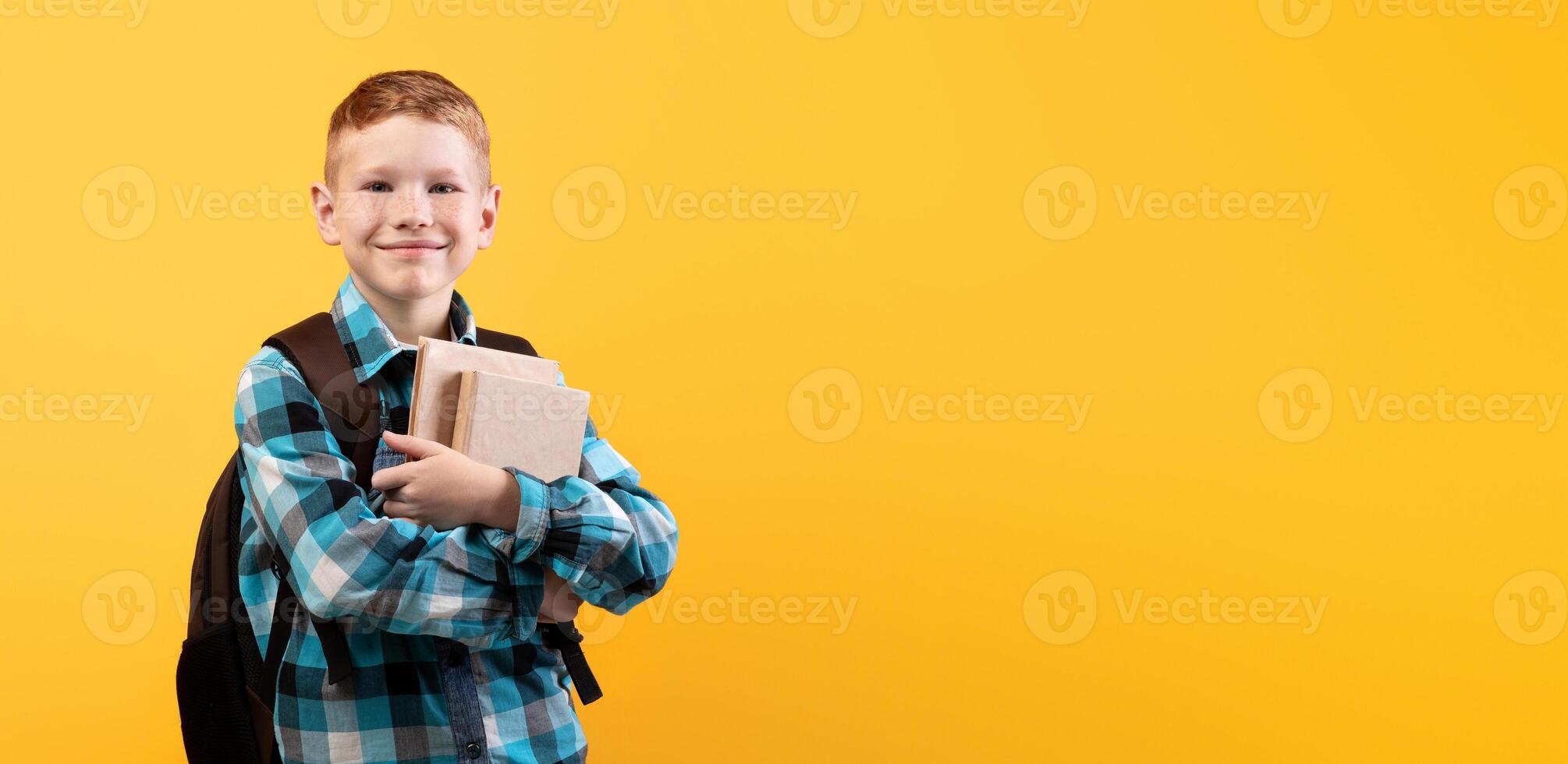 Cheerful schoolboy with backpack holding heap of books, yellow studio background, copy space. Happy boy going to school, holding books, enjoying his school days. Kids education concept photo