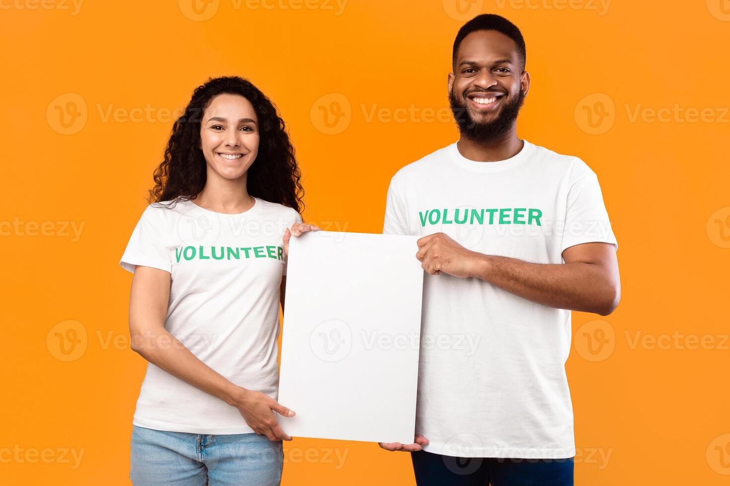 Two Diverse Volunteers Holding Empty Paper Poster For Your Text, Smiling To Camera Standing Over Yellow Studio Background. Volunteering Work, Charity Advertisement Banner. Mockup photo