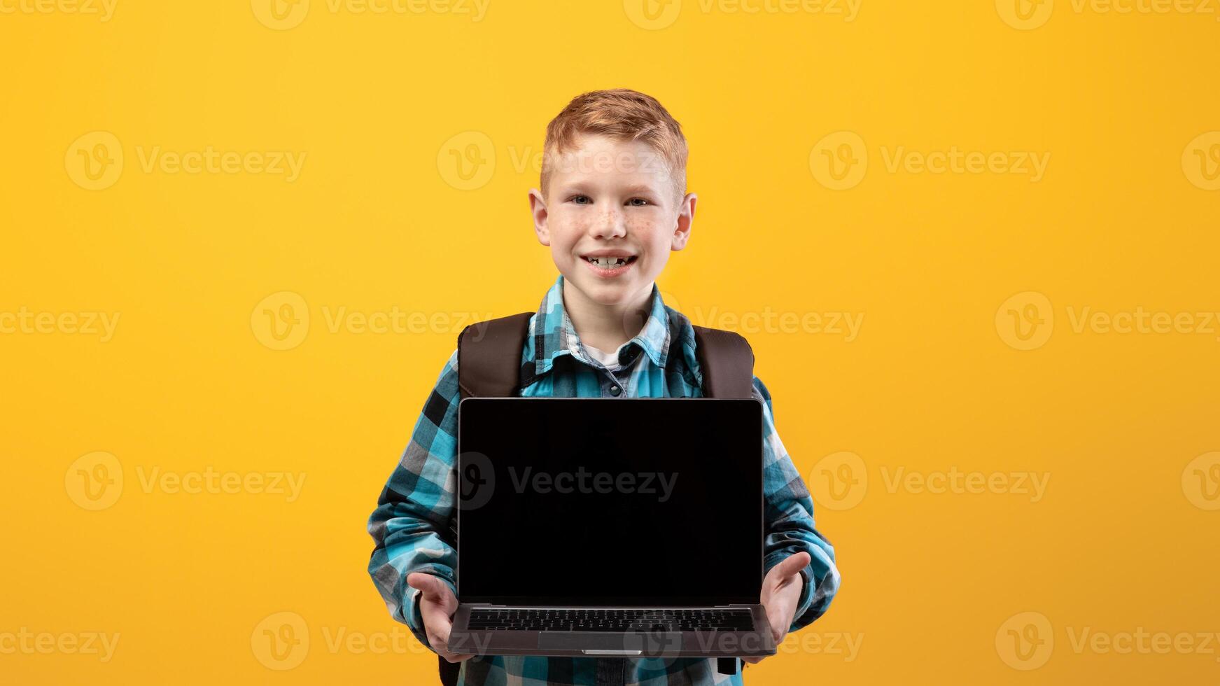 Cheerful ginger kid with schoolbag showing laptop with blank screen on yellow studio background, copy space. Happy schoolboy holding modern laptop, using technologies in education, mockup photo