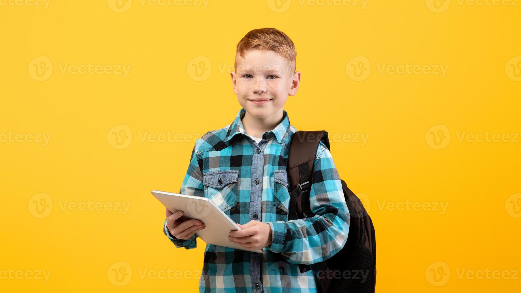 Technologies in school education. Smiling smart schoolboy with backpack holding digital tablet over yellow studio background, copy space. Cute kid using pad for studying, doing homework, websurfing photo