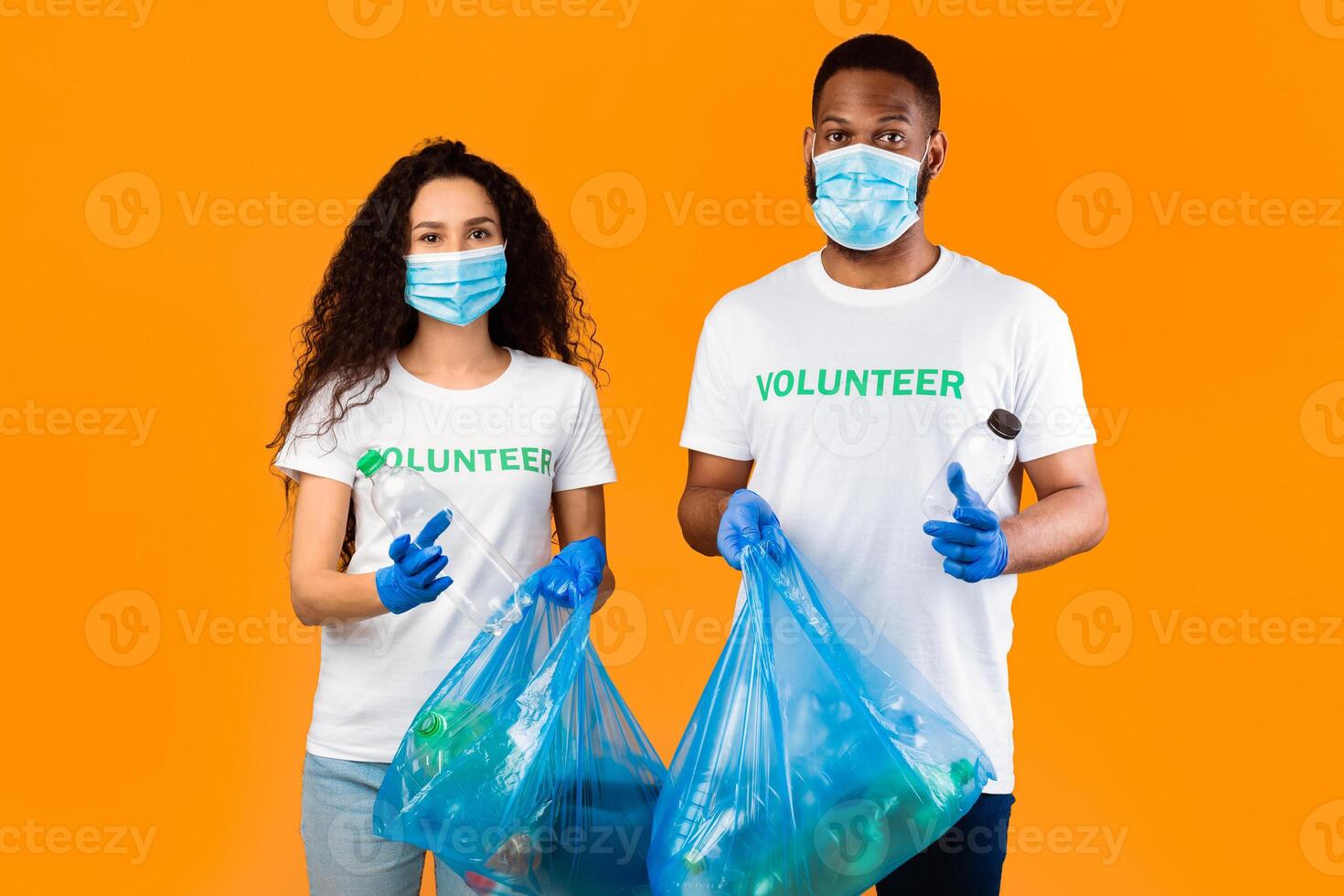 Two Multiethnic Volunteers Holding Plastic Bottles And Blue Garbage Bags With Litter Standing On Yellow Studio Background. Waste Sorting And Plastic Disposal. Keep Planet Clean Concept photo