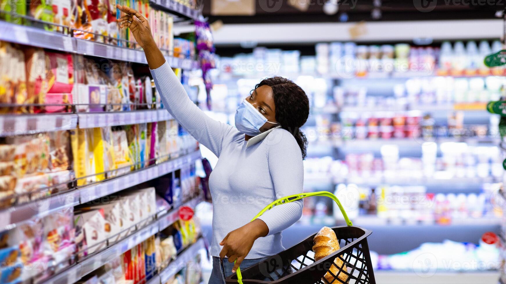 Black female customer in mask calling on mobile phone while shopping for food, reaching upper shelf at mall. Millennial African American woman buying groceries at supermarket during covid outbreak photo