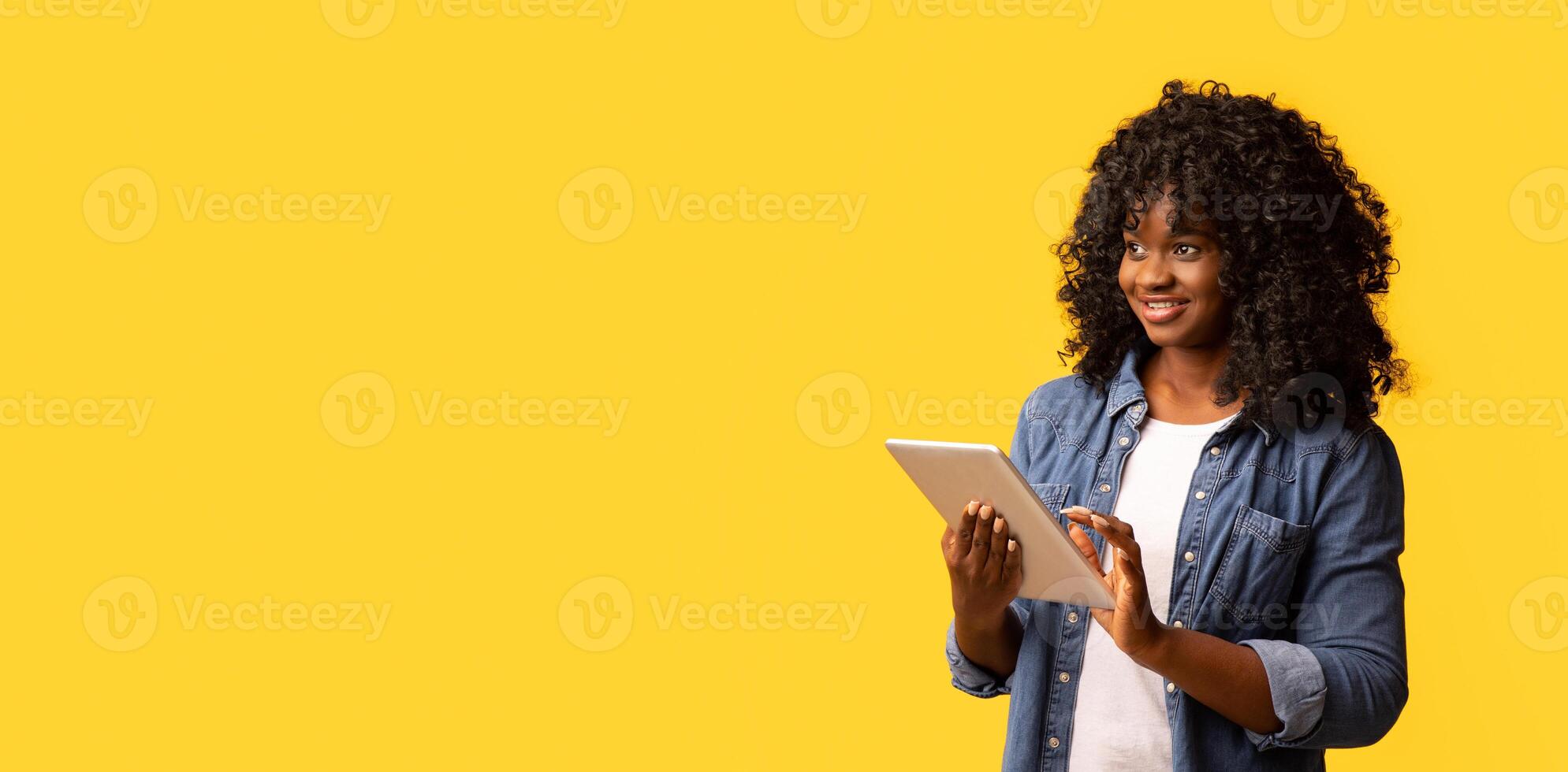 Joyful african american young woman with newest digital tablet looking at copy space on yellow studio background and smiling, panorama. Advertisement for mobile application concept photo