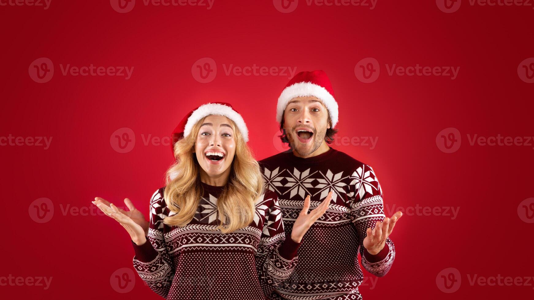 Excited millennial couple in Santa hats and Christmas sweaters on red background photo