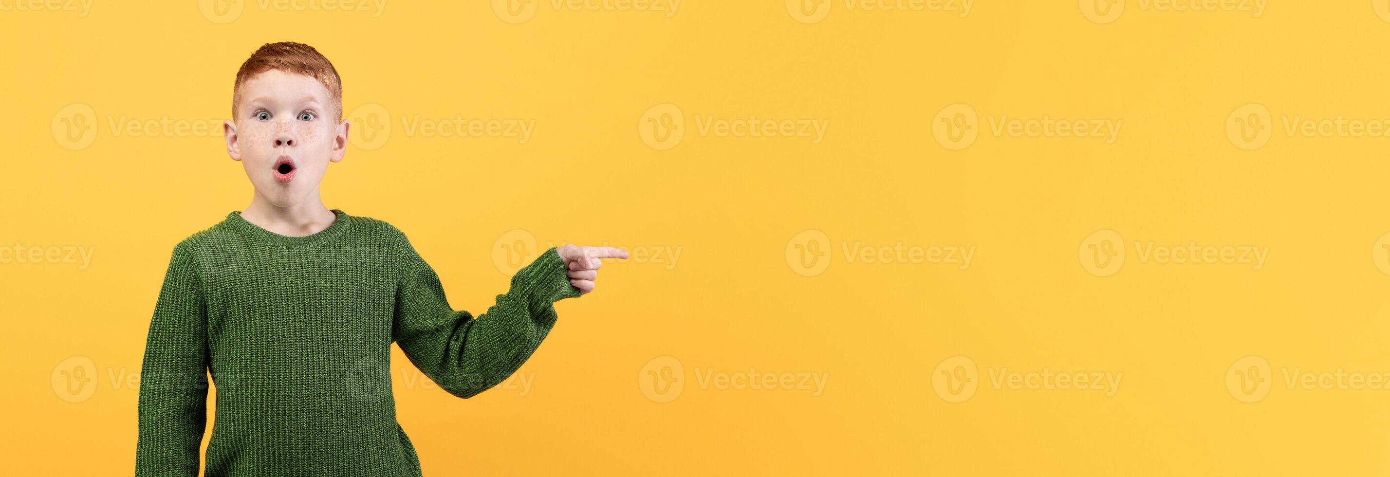 Excited ginger boy in green winter sweater pointing at copy space over yellow studio background, panorama. Shocked kid redhead guy showing amazing deal or announcement, winter sale concept photo