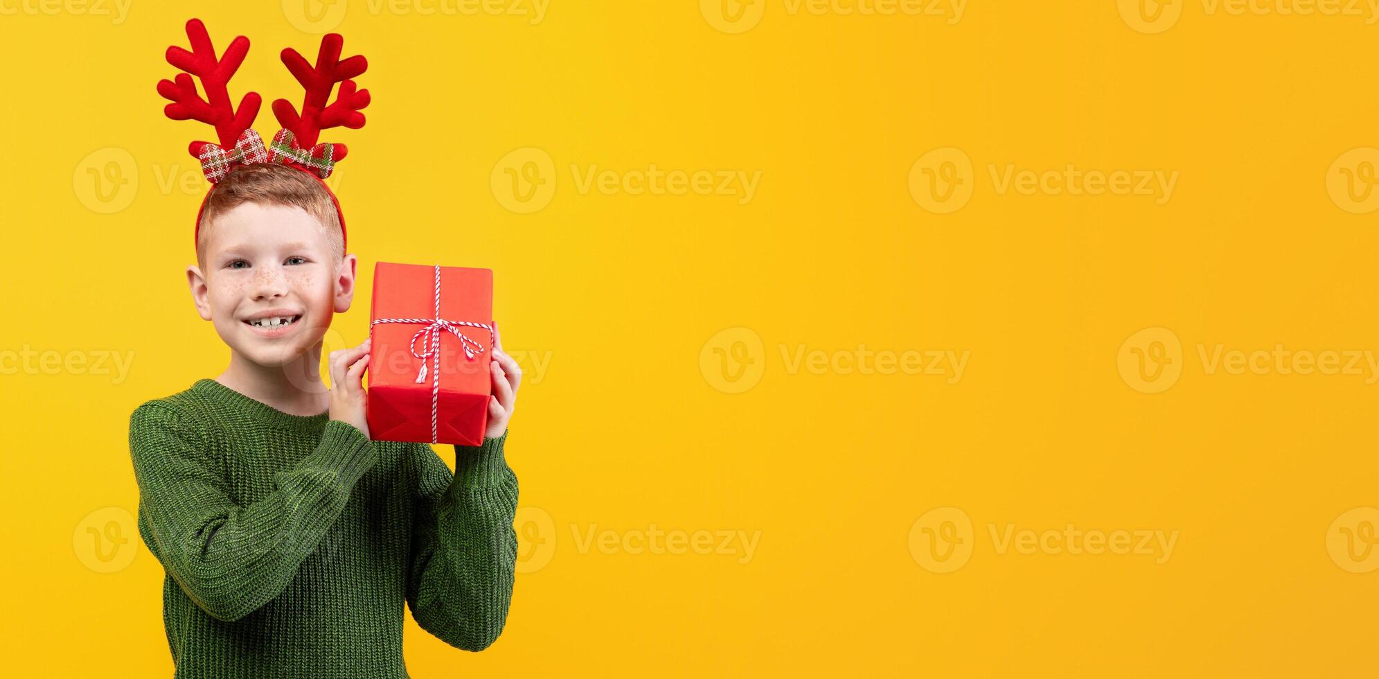 Happy kid with New Year gift over yellow studio background, copy space. Excited boy with deer accessory holding xmas present next to ear, wondering what is inside. Winter holidays celebration photo