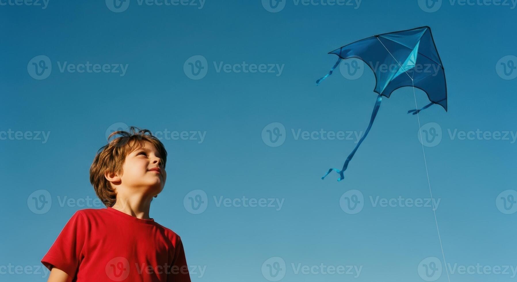 Young boy in red shirt looking up at a blue kite flying high in a clear sky photo