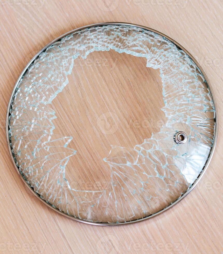damaged glass lid with metal rim, shattered with pieces missing, is sitting on light colored wooden surface photo