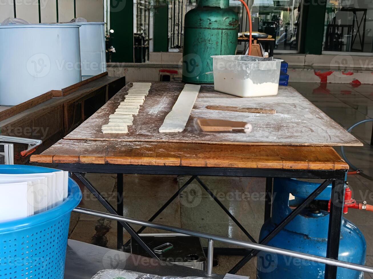 Rustic wooden table with dough and baking tools in bakery or kitchen setting, with large gas tank and containers in background, showing workspace for food preparation and baking photo