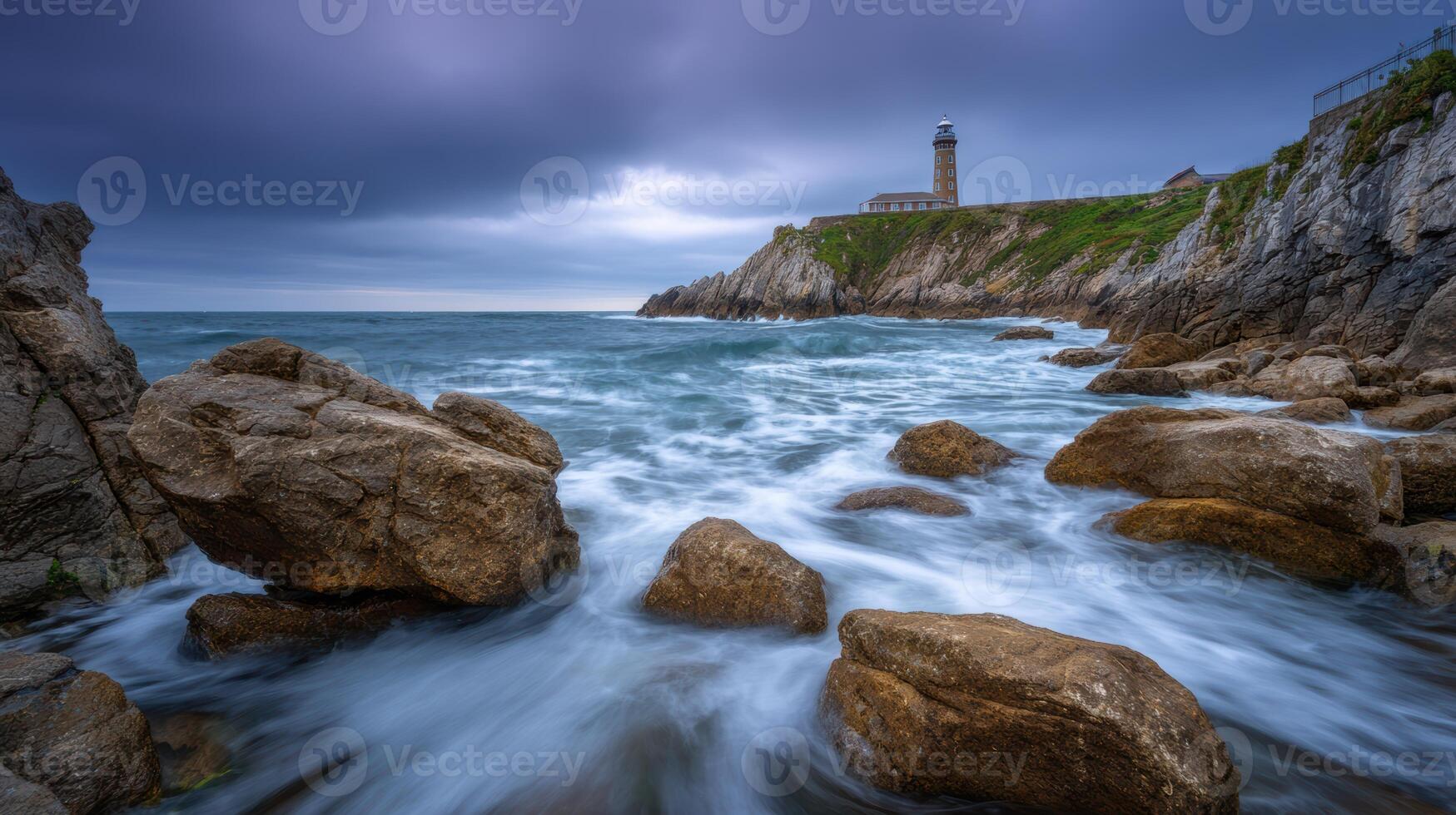 Rugged coastline scenery features a historic lighthouse perched atop a rocky cliff overlooking dynamic ocean waves under a dramatic sky photo