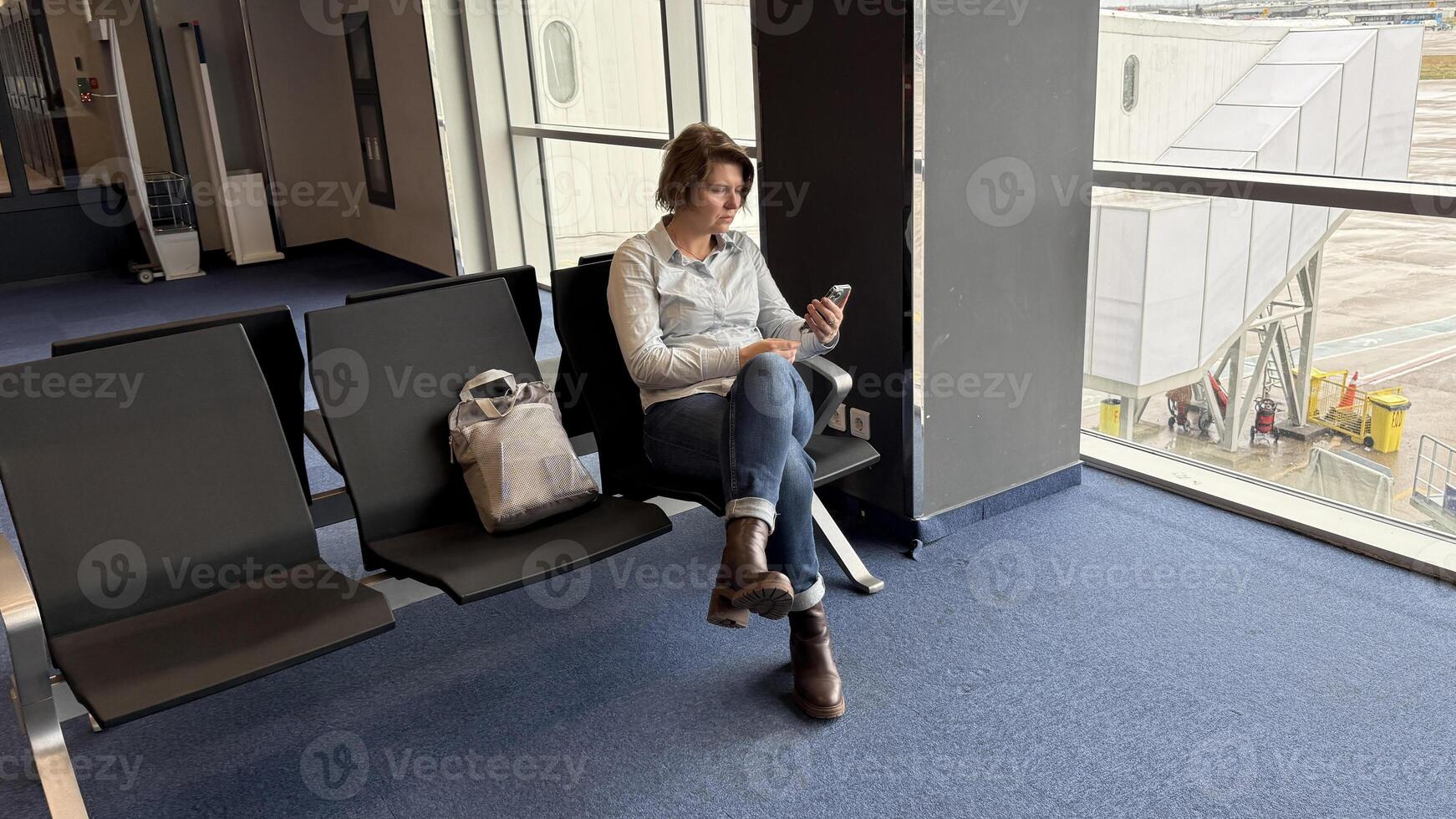 Seated alone on blue airport carpet, a Caucasian woman curiously anticipates Lunar New Year celebrations, phone in hand photo