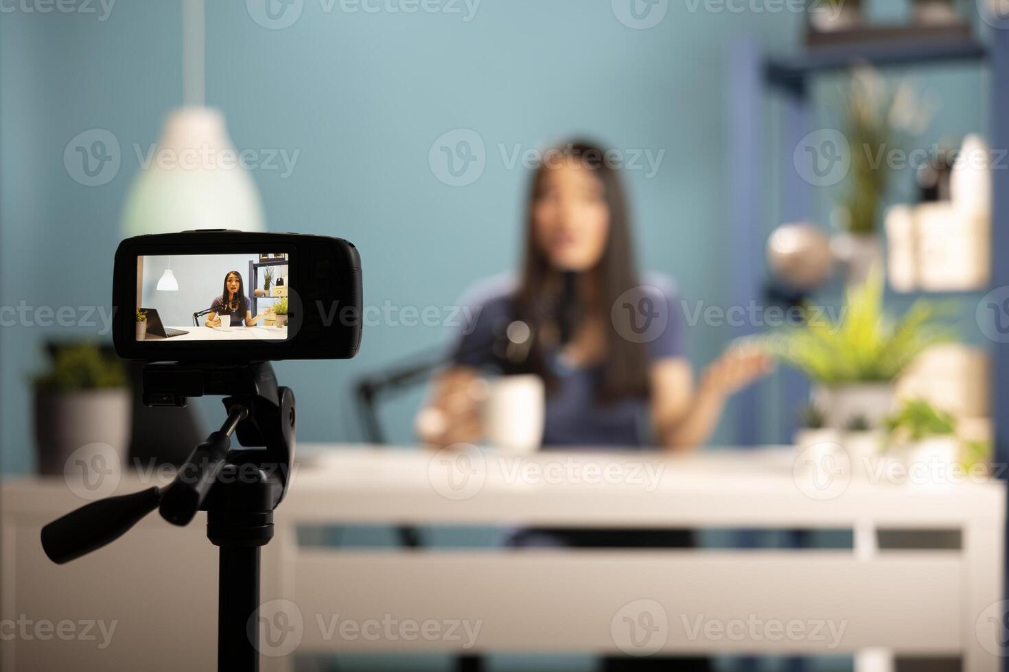 Asian woman blogger seated at desk in a professional studio setting, holding a cup of coffee and speaking into a microphone. The image emphasizes digital content creation and filming equipment. photo