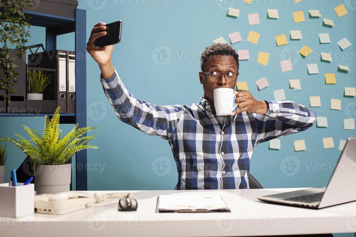 Black man seated at his office desk, drinking coffee and recording himself on his smartphone, documenting his workday. African american business advisor taking a selfie and enjoying a cup of tea. photo