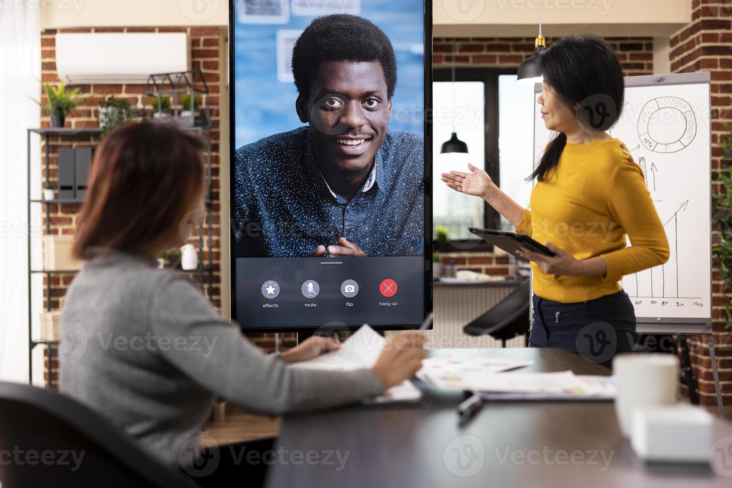 Female consultant stands with tablet, and coworker sits at desk, presenting company strategy ideas to male client during virtual meeting. Businesswomen engaged in videoconference call with black man. photo