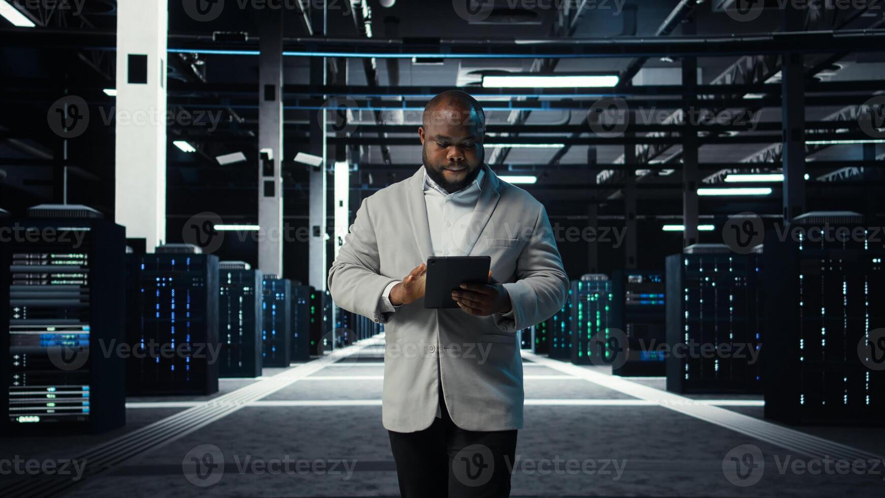 IT professional looking over data center checkup checklist paperwork on tablet. African american man walking in server farm, reading documentation files on device, camera A photo