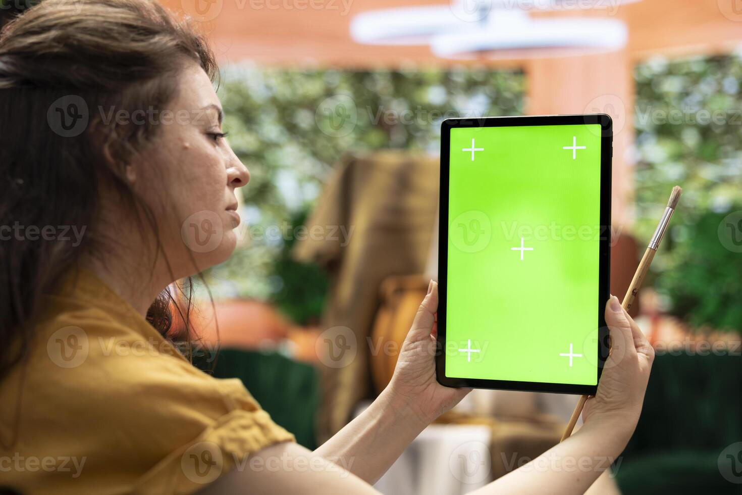 Woman painter using a tablet with copy space chroma key display sitting in her art and craft workshop, cozy creativity atelier. Artist enjoying masterpiece on canvas, artistic hobby. photo