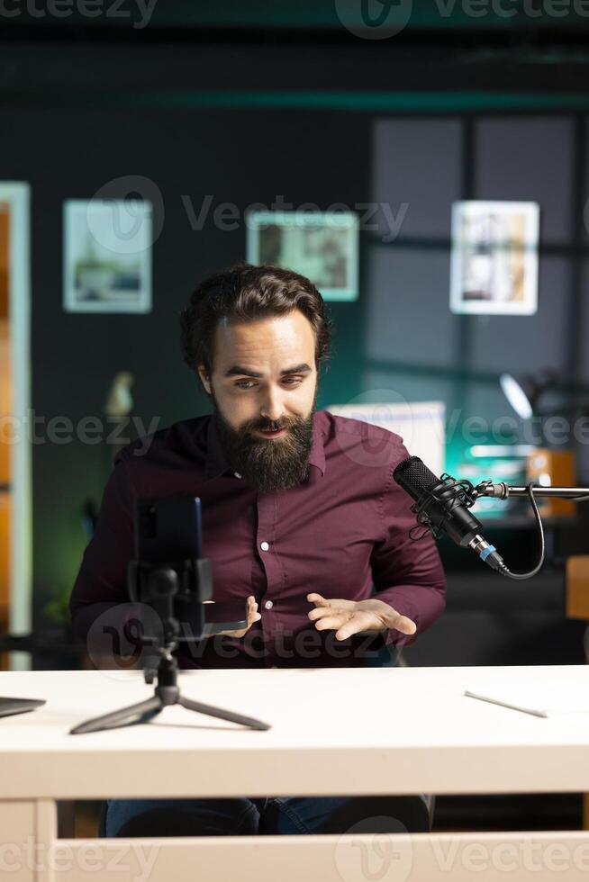 Man in studio endorsing smartphone from brand sponsoring , talking in professional microphone. Tech specialist does influencer marketing by promoting mobile phone, recording with high quality mic photo