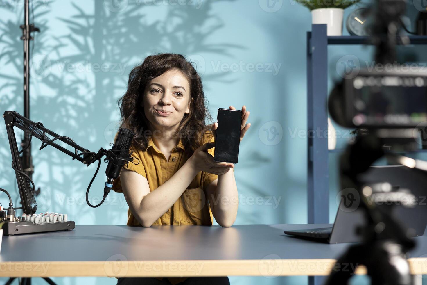 Cheerful entertainer presenting mobile phone from brand sponsoring , urging viewers to purchase it. Smiling woman does influencer marketing by advertising smartphone to audience photo
