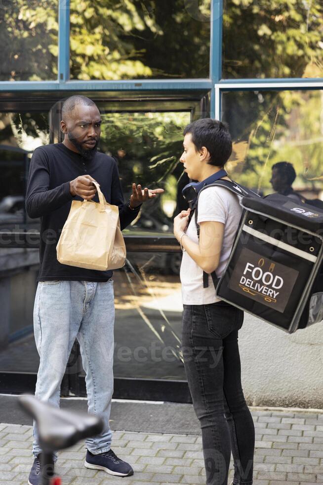 Black male client holding a takeout package with the wrong order, confronting the delivery worker about a shipping problem. The incorrect meal causes tension during failed courier service. photo