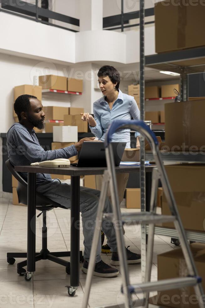 Diverse team at desk discussing order processing to guarantee distribution in a tightly packed storage room filled with products and boxes. Organized structure of a small scale business. photo
