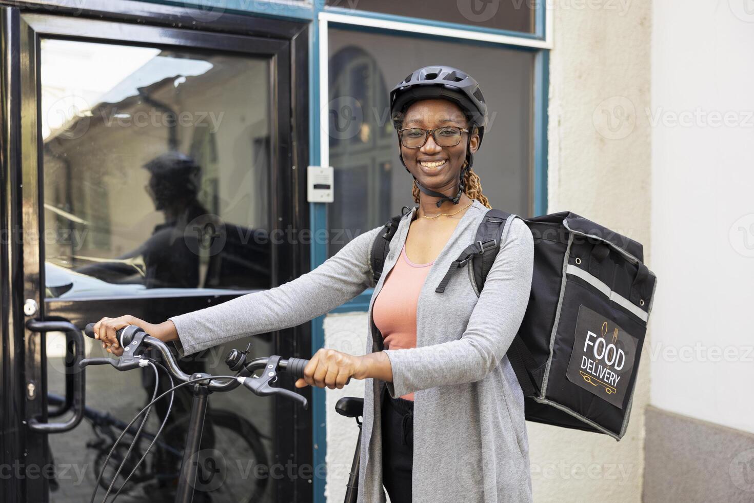 Portrait of smiling black woman working as a takeaway courier on the bike, riding with a helmet and carrying her backpack. Takeout employee delivering fast food or other meal orders. photo