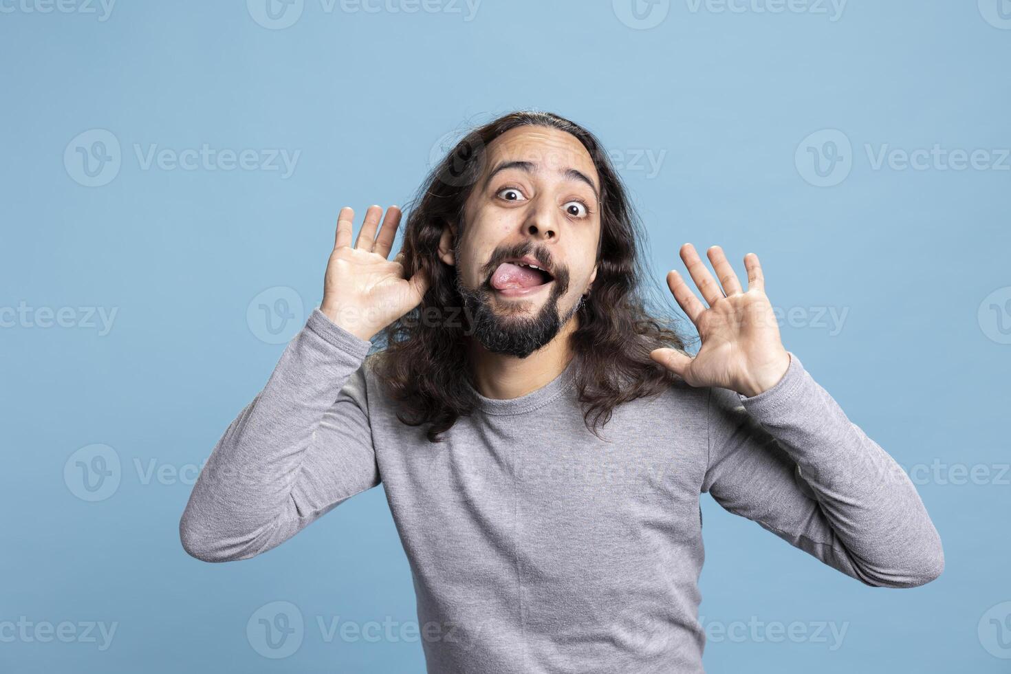 Playful young man with a long hair and beard making a goofy face in the studio. Carefree guy radiates humor and joy, capturing a lighthearted moment full of laughter and casual personality. photo