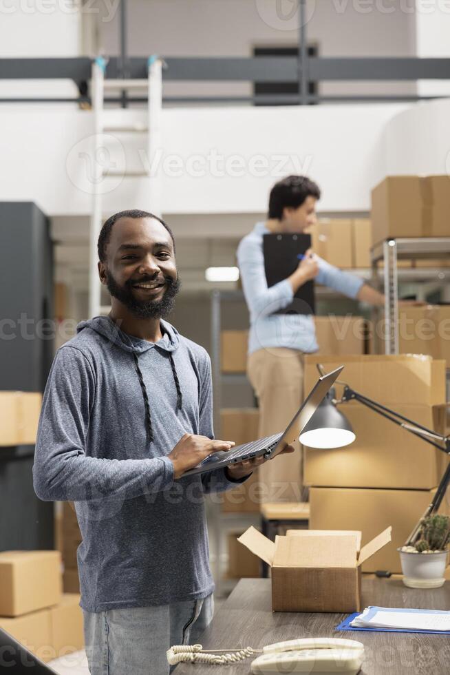 Warehouse employee using laptop to coordinate orders and ensure the distribution activity, small business logistics process. Preparing taped boxes across the organized supply room. photo