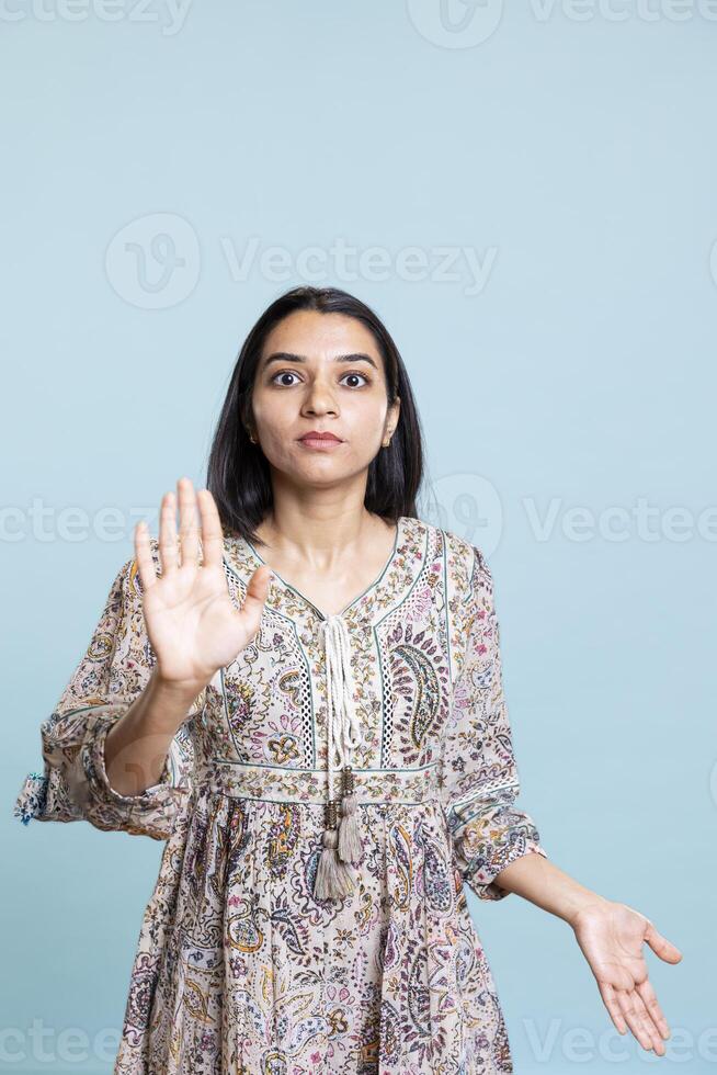 Focused firm indian woman showing stop hand symbol and complaining in studio. Authoritative person doing firm rejection symbol with her palm, having enough against blue background. photo