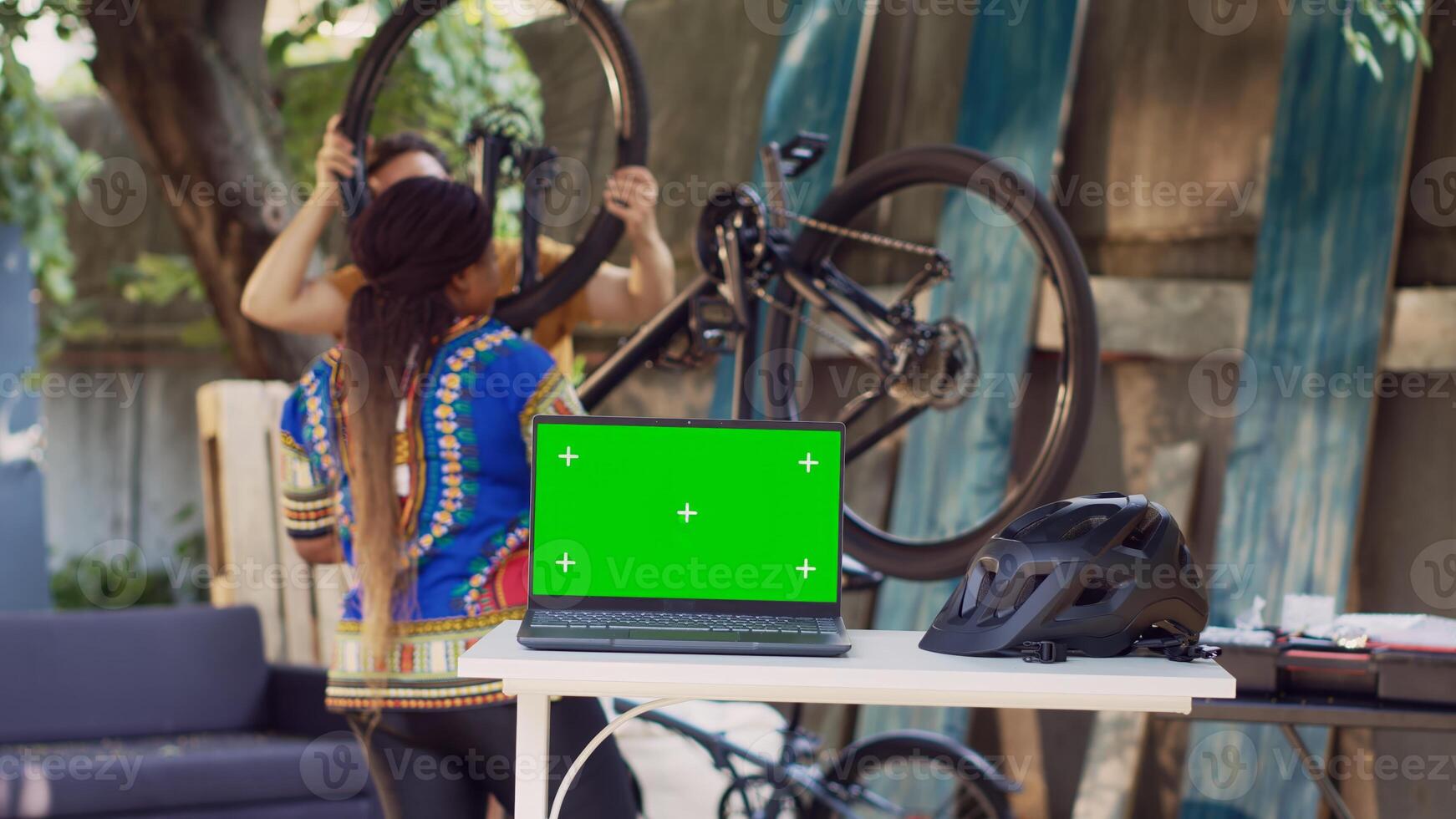 Laptop showing greenscreen while healthy and active interracial pair fixes bicycle components. Minicomputer with isolated copyspace screen on table while couple does bike maintenance at home yard. photo