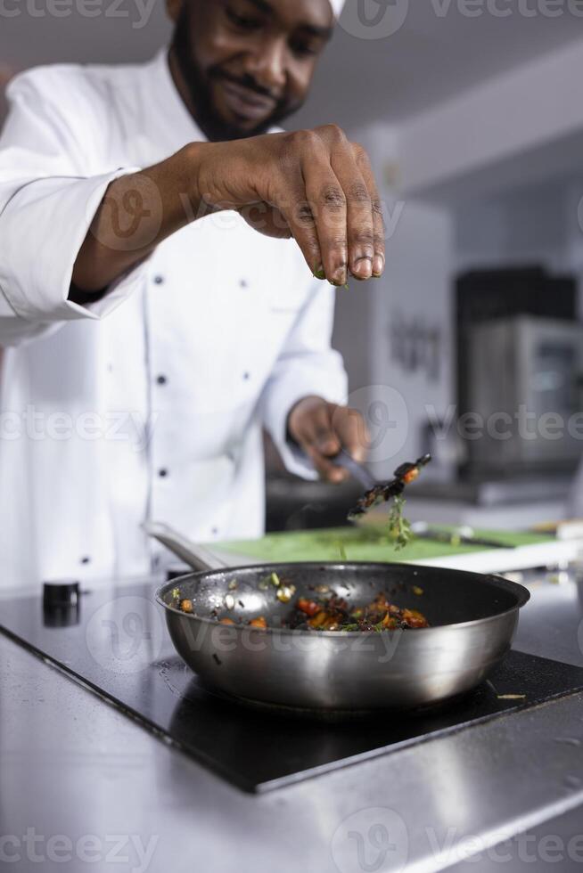 African american expert in a kitchen prepares a dish with fresh basil and parsley, showcasing culinary expertise in fine dining and food prep. Young person chef sprinkles rosemary leaves. photo